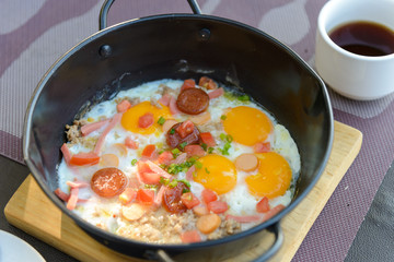 Top view of pan-fried egg for breakfast topping with pork, sliced ham, sausage, tomato and spring onion in a metal bowl