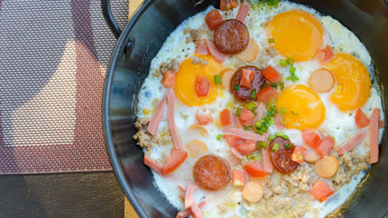 Top view of pan-fried egg for breakfast topping with pork, sliced ham, sausage, tomato and spring onion in a metal bowl