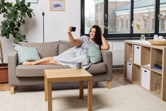 people, technology and leisure concept - happy young woman lying on sofa and taking selfie with smartphone at home