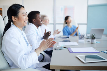 Fototapeta premium Side view at multi-ethnic group of doctors applauding while sitting at table during medical conference, focus on smiling Asian woman in foreground, copy space