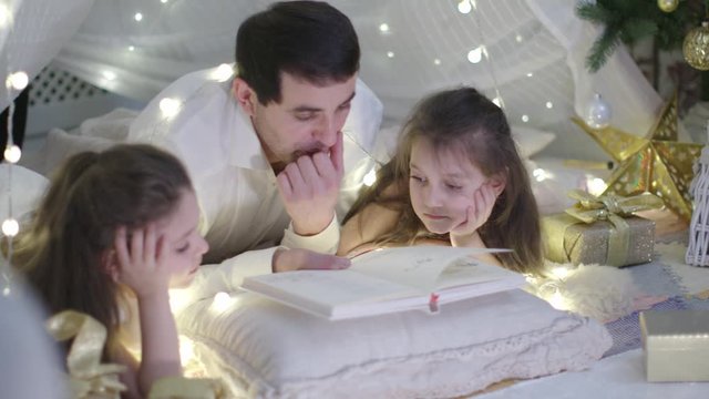 Father in White Shirt and Two Little Girls in a White Dresses Lies on the Carpet and Reading Book Together in Christmas Decoration. New Year Family Holiday. The Concept of a Parenthood, Family.