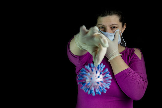A dark room. In it, a girl with dark hair with one hand holds a medical mask at her mouth. On the other hand is a model of the coronavir molecule