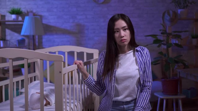 Tired sad young asian mom rocks a baby cot