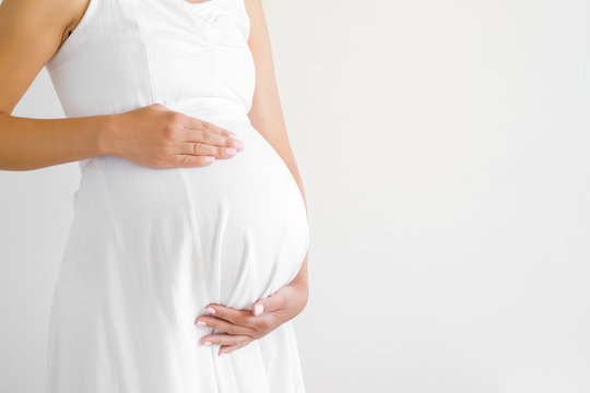 Young pregnant woman in white dress touching big belly with hands. Baby expectation. Pregnancy time - 37 weeks. Closeup. Empty place for text on gray background. Side view.
