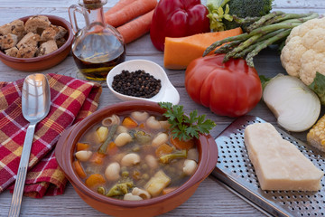Closeup view of a wooden table with a fresh, ready-to-eat homemade vegetable soup. Raw vegetables on the wooden table, black pepper, olive oil, croutons and parmesan