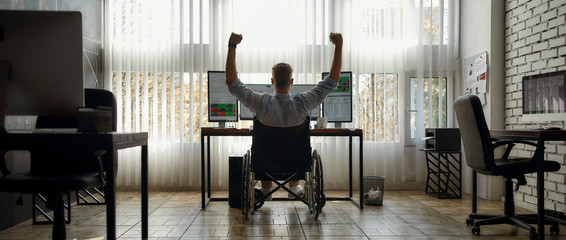 Winner. Back view of excited young male trader in a wheelchair looking at computer monitors and keeping arms raised while working in the modern office. Disability concept