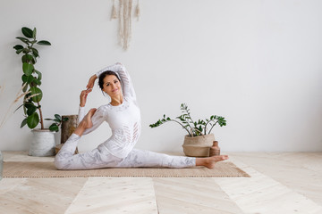 Slim pretty positive young brunette woman stretching in Monkey God exercise, Splits, Hanumanasana pose on mat on floor surrounded by houseplants on white wall. Advertising space. Yoga and pilates