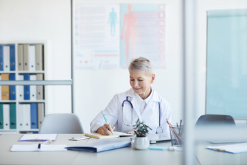 Portrait of mature female doctor writing notes and smiling while sitting at table in clinic interior, copy space