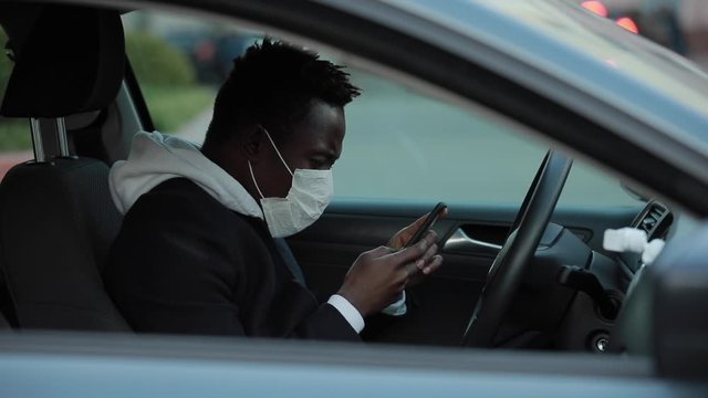African american man in surgical mask in a car reading news from smartphone about epidemic of virus covid-19