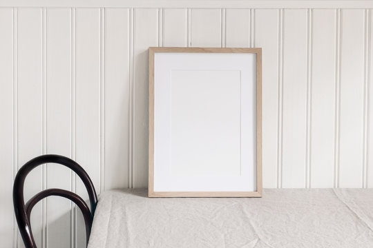 Empty wooden frame mockup on beige linen tablecloth background. White beadboard wainscot wall paneling background. Artistic still life with chair. Scandinavian interior, home design. Art concept.
