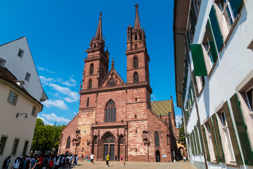 A group of tourists are waiting in front of the Basel Minster on a nice sunny day with blue sky. With its red sandstone walls, colourful roof tiles and twin towers it is a very famous landmark. 