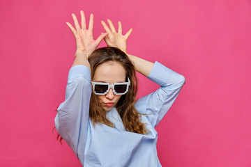 Stylish caucasian girl in a pale blue t-shirt and 8-bit glasses and makes bunny ears with his hands on a pink background.