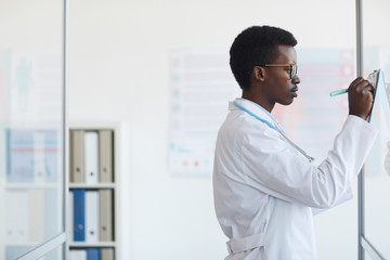 Naklejka na ściany i meble Side view portrait of young African-American doctor writing on clipboard while standing by glass wall in med clinic, copy space