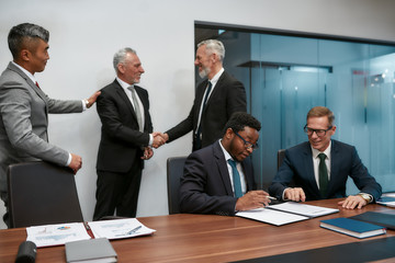Congratulations. Confident african businessman reading a contract and discussing something with his partner while two business people in formal wear shaking hands on the background
