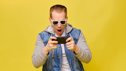 Stylish Caucasian man in a jeans on a yellow background with 8 bit glasses. Play with surprise on a smartphone.