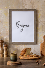 Stylish interior of kitchen space with wooden table, brown mock up photo frame, bread, cup of tea, basket and kitchen accessories in wabi sabi concept of home decor. 