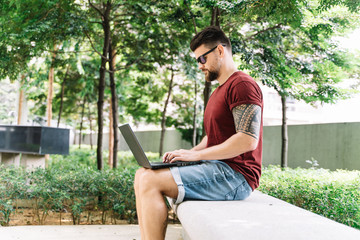 Fototapeta premium Man sitting on a bench in the middle of a park working remotely with his laptop