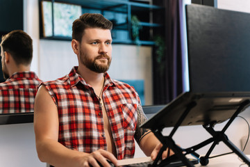 Man with a beard and a plaid sleeveless shirt working remotely in front of a laptop
