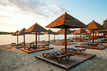 Evening sandy beach with brown wooden loungers and umbrellas. Empty rows resting places. Recreation calm lake in light of the low evening sun.