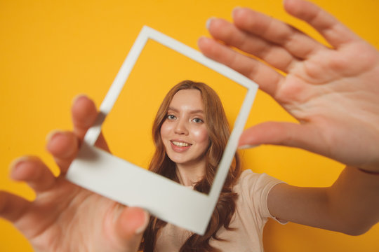 Young Smiling Woman Holding In Hands Photo Frame With Her Face Isolated On Yellow Background, Selfie Concept