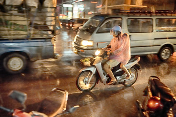 Parent with child hidden under plastic disposable cap riding scooter in city traffic at night under heavy tropical rain. High iso photograph.