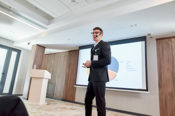 Mission Possible. Young male speaker with headset and laser pointer in suit presenting a chart on the screen while giving a talk at business meeting, economic forum
