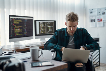 Portrait of young focused male web developer in a wheelchair writing program code on his laptop at his workplace with multiple computer screens on the background