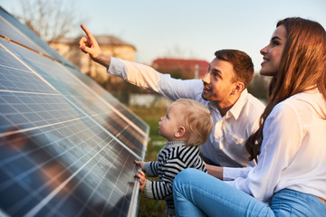 Man shows his family the solar panels on the plot near the house during a warm day. Young woman with a kid and a man in the sun rays look at the solar panels. - obrazy, fototapety, plakaty