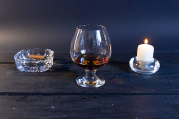 Brandy in glass on black background with cigar in ash tray and burning candle