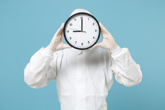 Man in white protective suit respirator mask hold in hand clock isolated on blue background studio. Epidemic pandemic new rapidly spreading coronavirus 2019-ncov from China medicine flu virus concept.