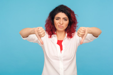 Dislike, bad review! Portrait of frustrated dissatisfied hipster woman with fancy red hair showing thumbs down, expressing disapproval, critique. indoor studio shot isolated on blue background