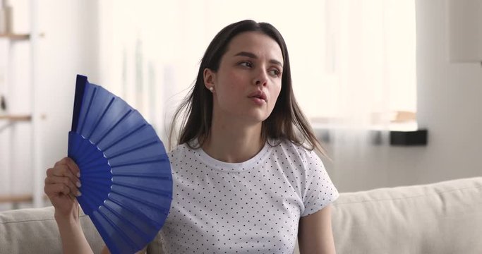 Unhappy young woman waving fan feeling uncomfortable hot and tired suffering from summer heat or humidity at home. Overheated lady sweating sitting on sofa in apartment. Broken air conditioner concept