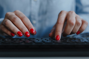 Closeup of woman's hands with red nails typing on a black keyboard wearing blue shirt at a...
