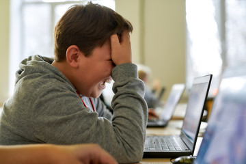 Tiresome process. Close up of caucasian schoolboy looking sad and desperate while using laptop together with other pupils during a lesson in modern smart school