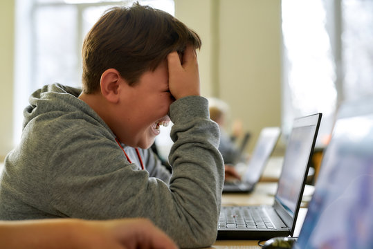 Tiresome process. Close up of caucasian schoolboy looking sad and desperate while using laptop together with other pupils during a lesson in modern smart school - Powered by Adobe