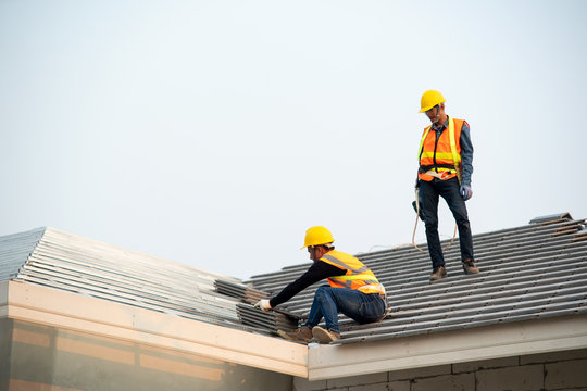 Roofer Worker In Protective Uniform Wear And Gloves,Using Air Or Pneumatic Nail Gun And Installing Concrete Roof Tile On Top Of The New Roof,Concept Of Residential Building Under Construction.