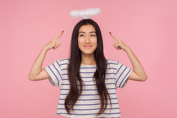 Obraz na płótnie Canvas Portrait of shy modest asian girl with long brunette hair in striped t-shirt pointing at angelic halo over her head, showing nimbus and timidly smiling. indoor studio shot isolated on pink background