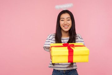 Charity and donations. Portrait of angelic asian girl with halo over head giving gift box to camera and modestly smiling, expressing kindness, generosity, care. studio shot isolated on pink background