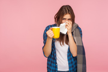 Treatment of seasonal flu. Sick girl wrapped in scarf blowing runny nose, sneezing in napkin and holding tea cup, treating influenza, cough and fever. indoor studio shot isolated on pink background © khosrork