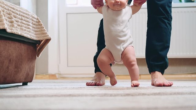 Little baby boy walking barefoot with help of father in home. Baby first steps