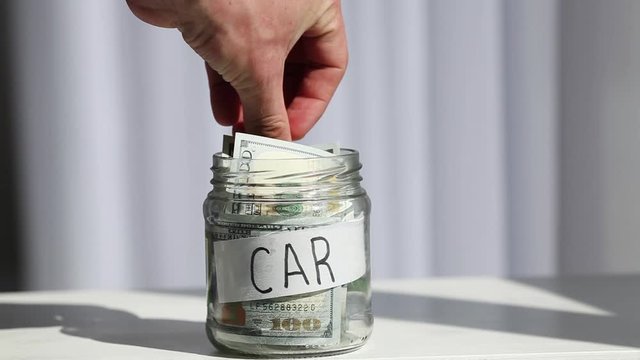 Male hand puts 100 dollars in a glass jar with the text car. Concept of saving money.