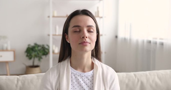 Calm young woman meditating with eyes closed at home alone. Happy mindful lady doing yoga breathing exercise relaxing on sofa. Healthy girl feels peace of mind, no stress concept. Close up view