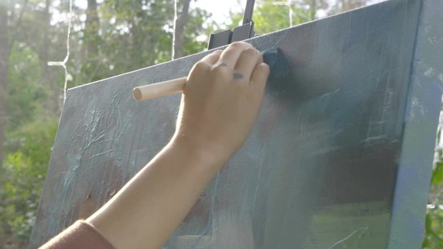 Female artist uses brush to dab top of abstract multicolored painting on canvas outside during the day, slow motion