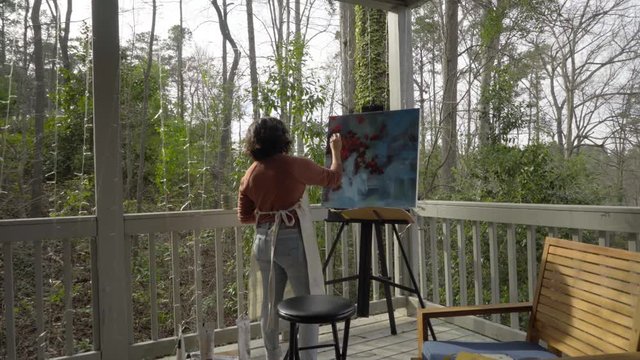 Young female professional painter works outside on multicolored abstract painting with brush during sunset, camera dolly left