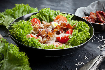 closeup of fresh Caesar salad with dried tomatoes, parmesan cheese, eggs and croutons in black bowl on dark background