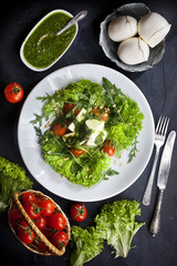kitchen table with fresh mozzarella salad, lettuce and arugula leaves, pesto sauce, fresh cherry tomatoes in a basket, flatlay