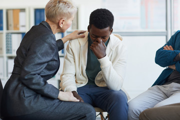 Portrait of young African-American man crying while sitting on chair in support group circle, copy...
