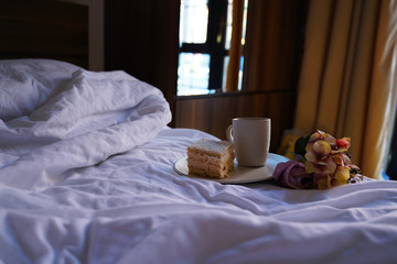 Breakfast in the bed. On the wite sheets the cup of coffe, flowers and the plate with cake