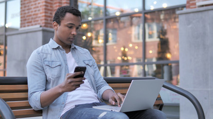 African Man Using Smartphone and Laptop, Sitting on Bench