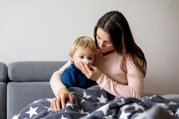Mother holding sick child, lying together on a couch at home with fever and running nose